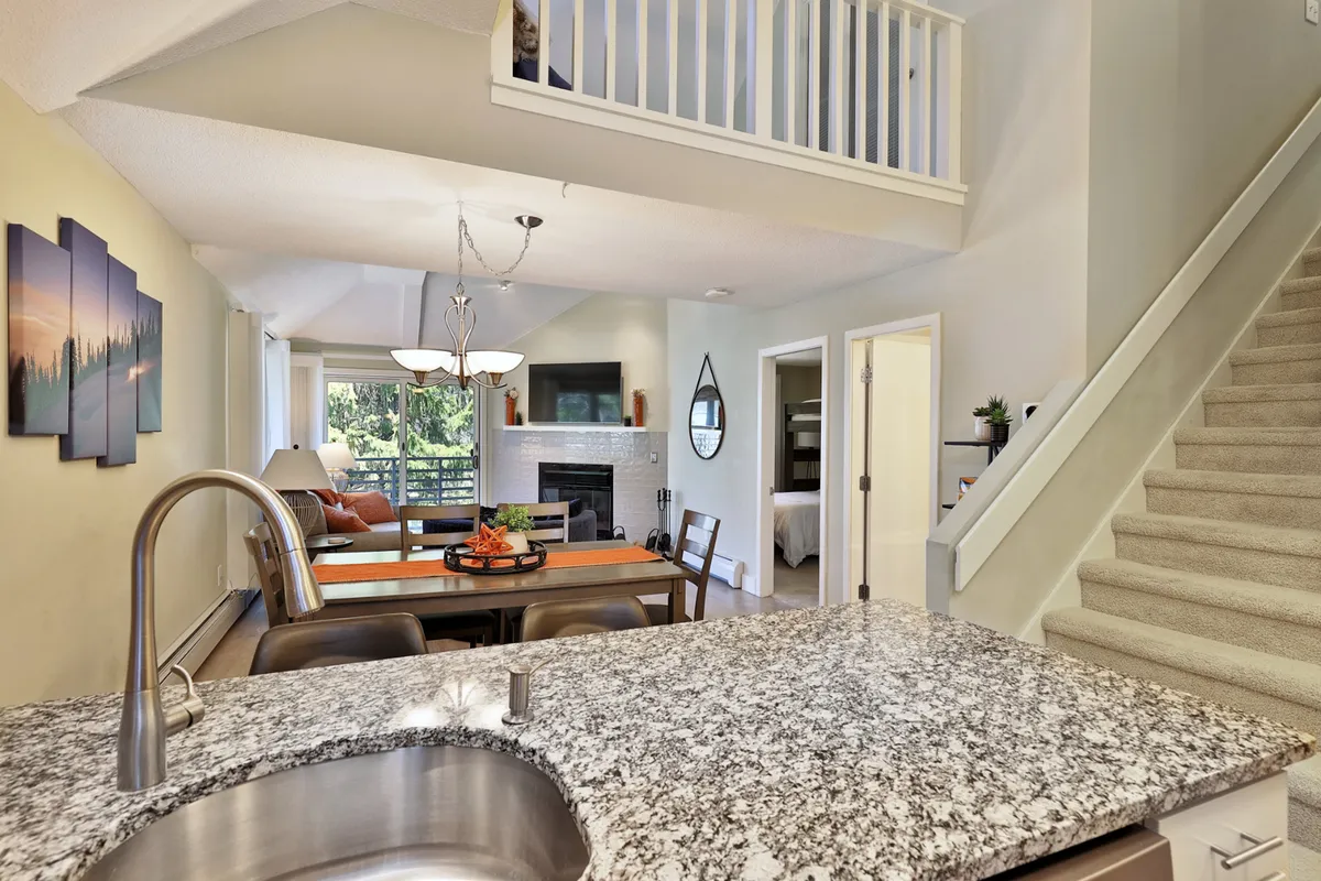 This image captures the heart of the open-concept living space, featuring a granite kitchen countertop, dining table, fireplace, staircase, and expansive windows letting in natural daylight. The kitchen and dining area are seamlessly connected, while the living area is centered around the fireplace and comfortable seating.