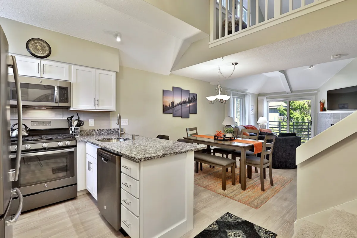 A view of the kitchen and dining area showcasing a granite countertop, dining table with chairs, artwork on the wall, and large windows allowing plenty of daylight. The setting is clearly designed for socializing, shared meals, and lively conversation.