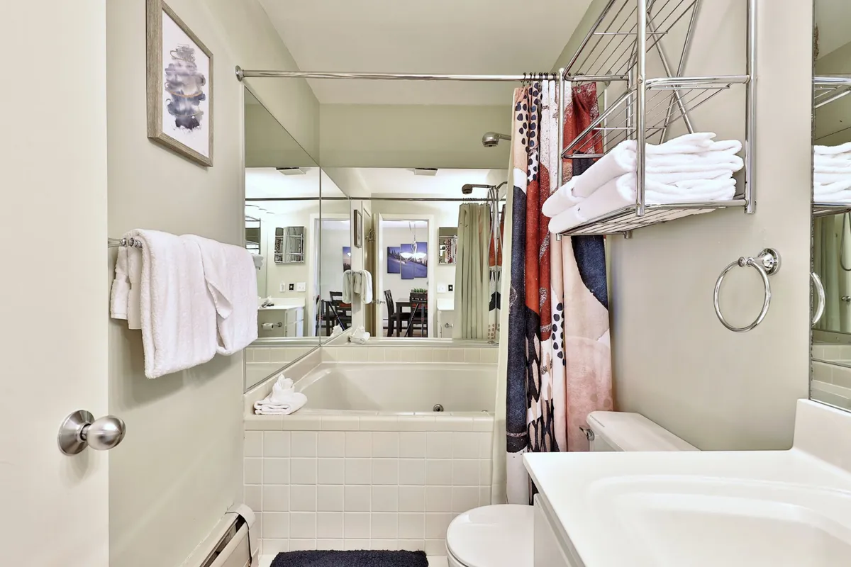 A well-lit bathroom scene featuring a bathtub, organized towel rack, colorful shower curtain, and mirror reflections. The space exudes freshness and order, with thoughtful touches such as accessible towels and vibrant decor, inviting guests to slow down and indulge in self-care routines.