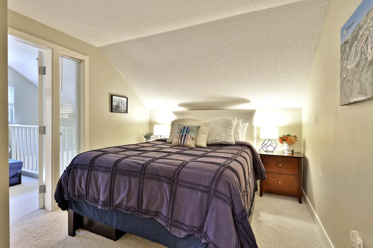 A restful bedroom with a plush queen bed, nightstand, lamps, and decorative pillows. The inviting bedding and tranquil setting evoke a sense of privacy and comfort, aligning with the description of the upstairs loft primary bedroom's serene ambiance.