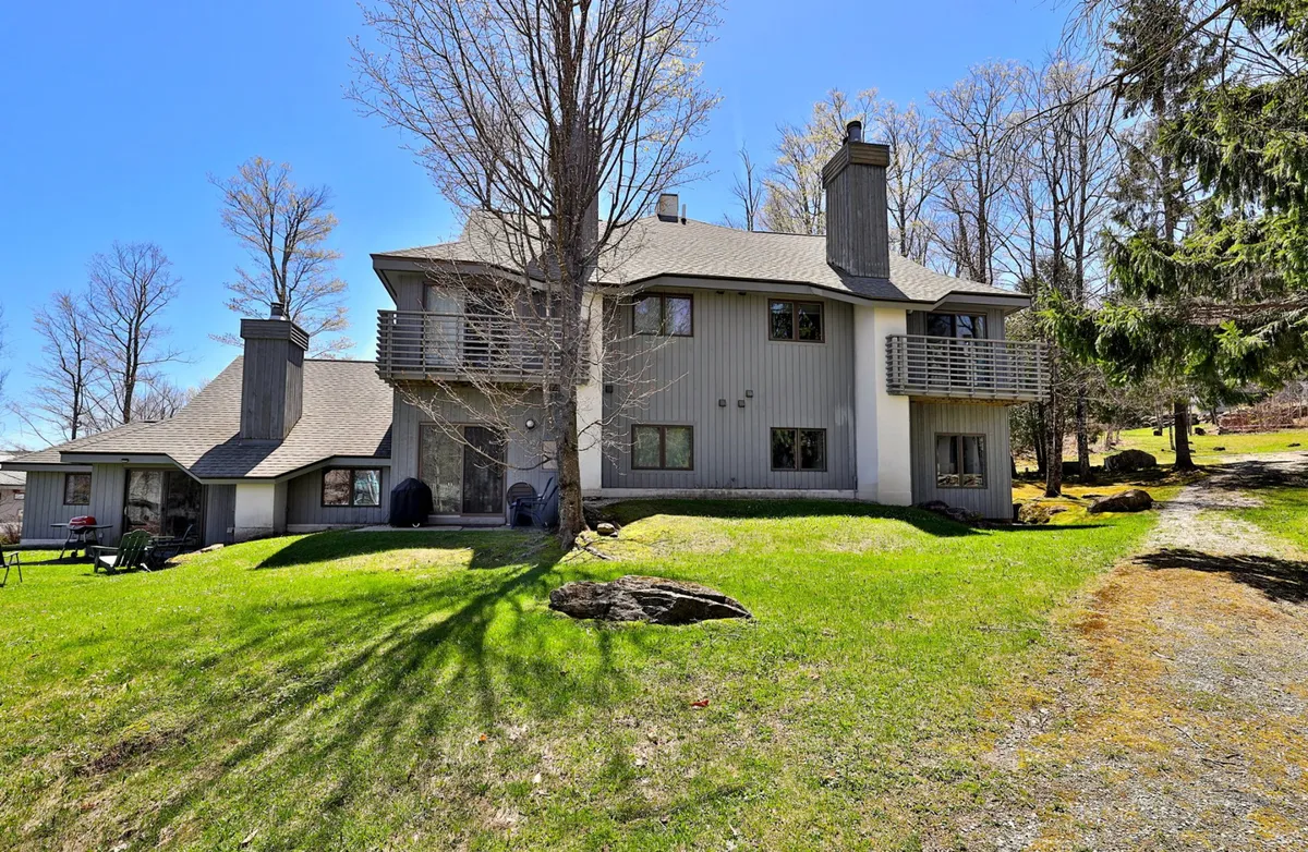 This exterior shot captures a welcoming two-story house surrounded by large trees and a green lawn. A balcony overlooks the outdoor area, emphasizing space for gatherings and relaxation. The scene is vibrant and sunlit, suggesting both summer fun and winter coziness at the doorstep.