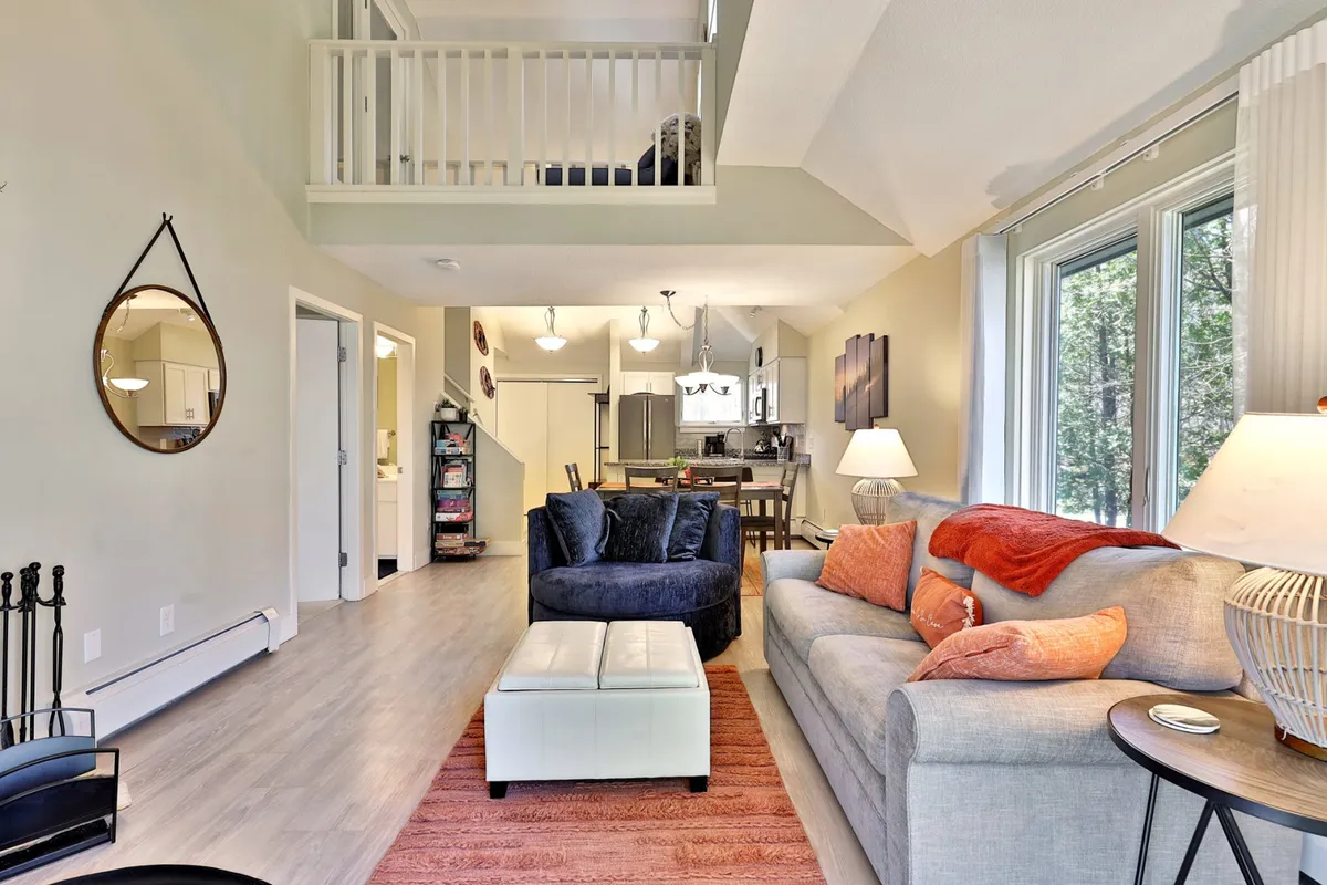 A comfortable gray sofa with colorful cushions sits in the foreground, a blue armchair and white ottoman complete the seating area, while the dining area and natural light–filled windows are visible in the background, reinforcing the open-plan layout.