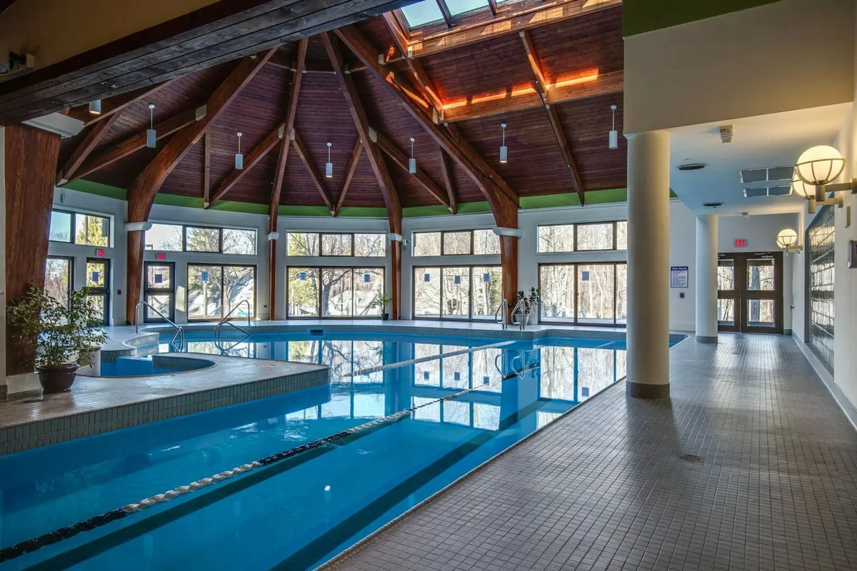This image showcases a serene indoor swimming pool accented by skylights, wooden beams, and large windows, allowing natural light to flood the space. Guests are seen swimming, lounging by the pool, and socializing, evoking a calm and rejuvenating mood.