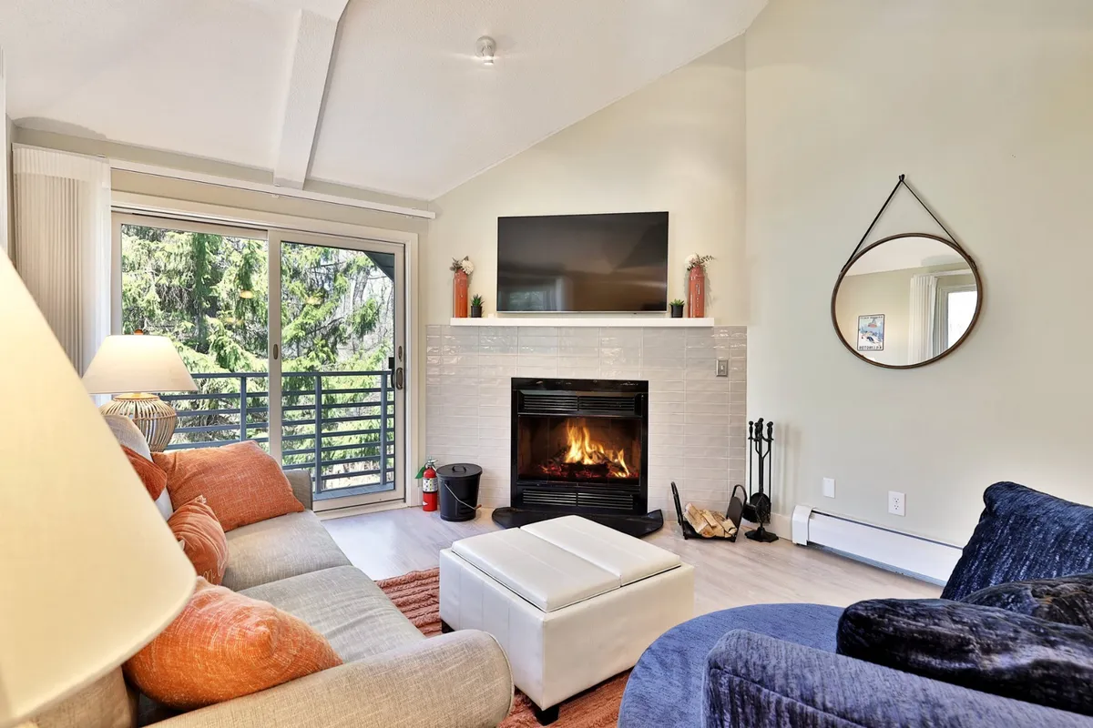 A cozy living room centers on a wood-burning fireplace, plush sofa, and large windows that frame the changing seasons outside. The scene feels inviting and relaxing, ideal for unwinding after a day of outdoor adventure.