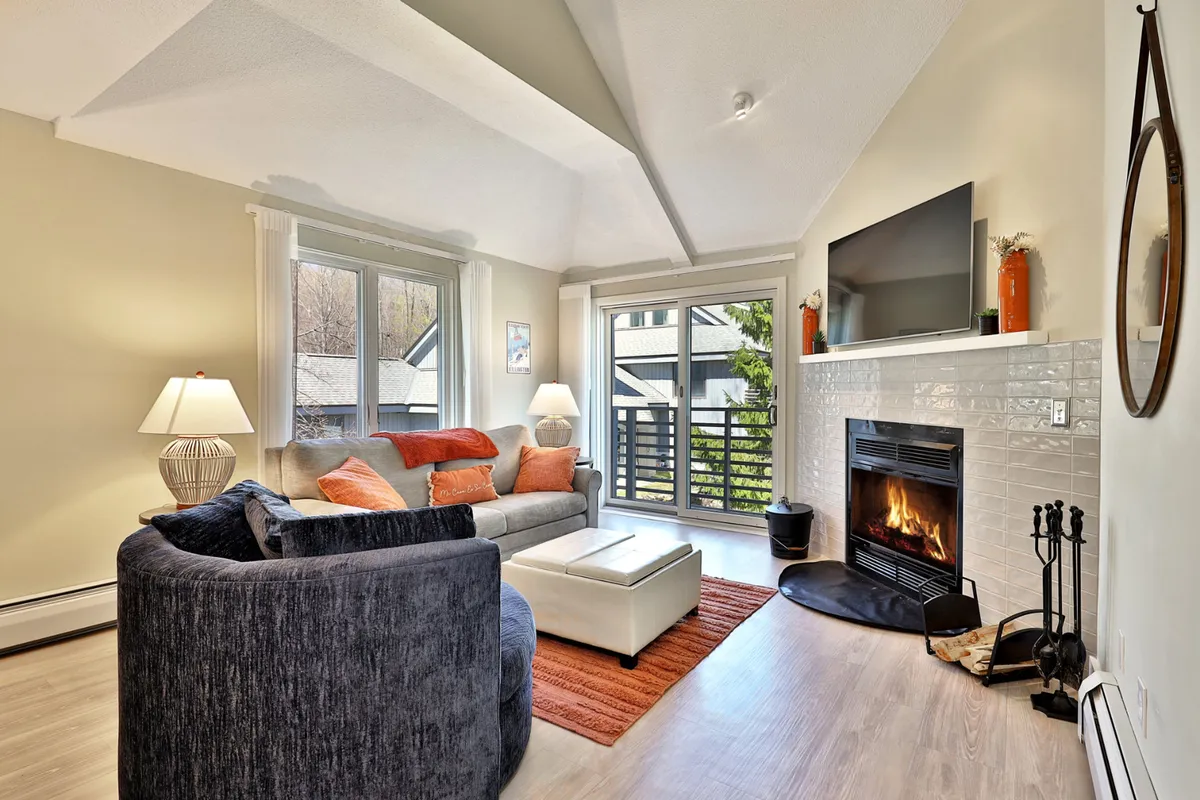 The image showcases a stylish condo living area with a cozy fireplace, inviting sofa adorned with plush decorative pillows, and expansive windows that fill the room with natural light. The scene evokes a sense of warmth and relaxation, perfect for unwinding after a day on the slopes or gathering with friends and family.