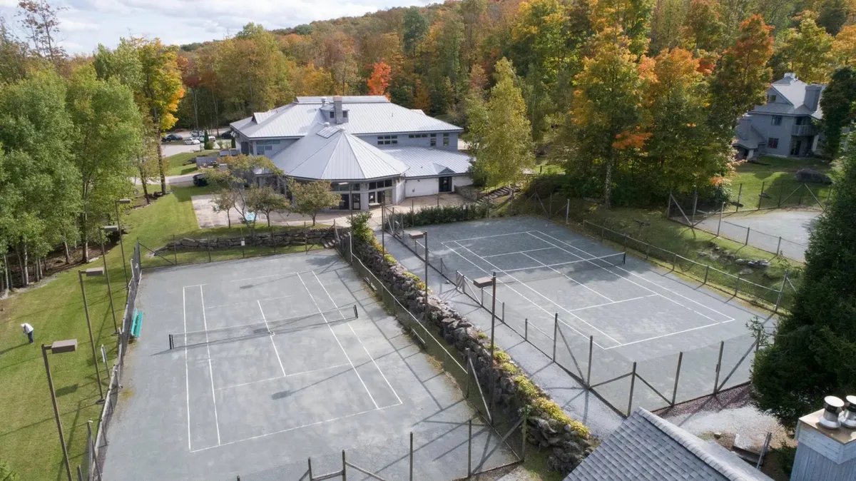 An exterior shot featuring well-maintained clay tennis courts adjacent to the main building, framed by lush trees. Guests can be seen enjoying tennis matches and outdoor activities, highlighting the vibrant, active lifestyle available.